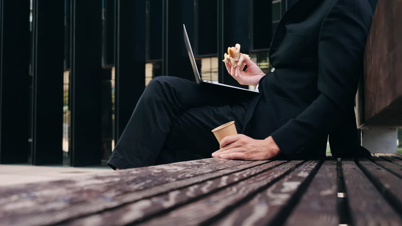 Man in suit working on laptop while eating sandwich on bench