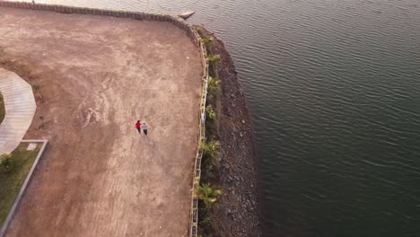 video de vista aérea de 4k de una pareja caminando con un perro pequeño shih tzu a lo largo de la orilla de un hermoso lago escénico de vacaciones o vacaciones durante el verano cerca de pune, india