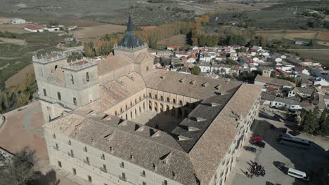 Vuelo-Con-Dron-En-El-Monasterio-De-Uclés-Con-Vista-Diagonal,-Aparece-Al-Fondo-El-Aparcamiento-Con-Vehículos-En-La-Entrada,-La-Iglesia,-El-Claustro-Y-El-Pueblo-De-Uclés