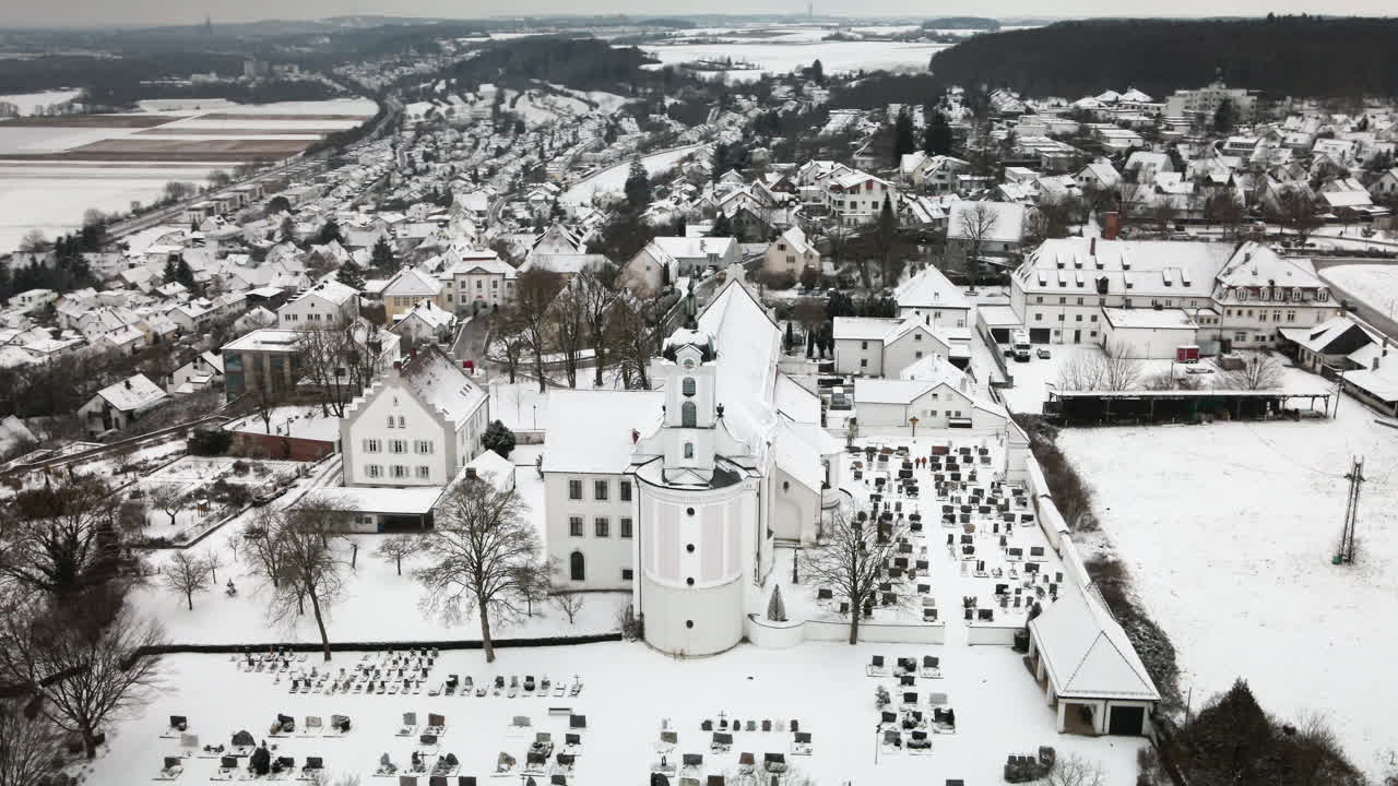 Dron kroužící doprava kolem zasněžené věže kostela v Elchingenu, hřbitova, střech vesnice a zasněžených polí. Ideální pro cestopisné dokumenty, reklamy a odhalení