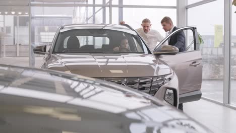 young car salesman showing to young couple new automobile at dealership salon.