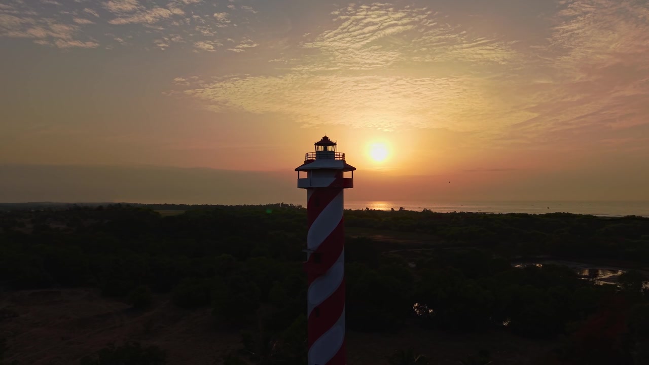 Ripresa aerea con drone che cattura un faro a strisce stagliato contro il sole nascente mentre una luce calda si diffonde sul Golfo del Bengala vicino a Chennai nel Tamil Nadu, India