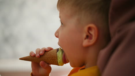 close up side view of young boy licking mint chocolate ice cream cone while holding it firmly in hand, sitting relaxed with soft blur white textured background