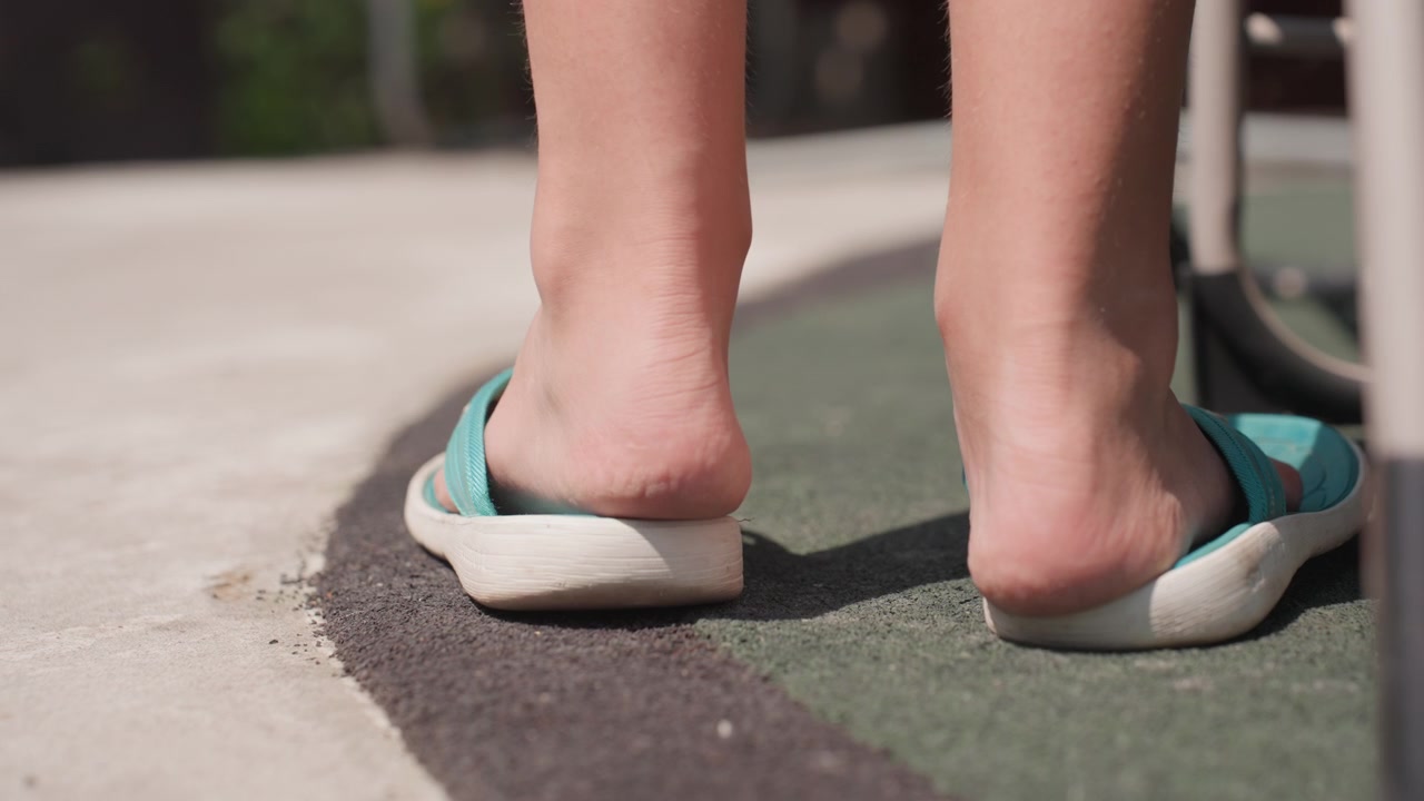 Pair of feet and turquoise flip flops on textured outdoor surface. Feet move into view pick up one sandal from ground then both sandals are worn as walking begins. scene features soft