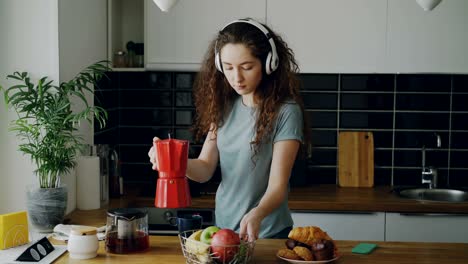 hermosa adolescente caucásica rizada con auriculares bailando preparando el desayuno, bebiendo café, poniendo la tetera roja, tomando y navegando en el teléfono inteligente