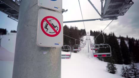 Abandoned-stopped-chairlift-in-an-empty-ski-resort,-sign-on-a-pole,-ski-slopes-deserted-during-Covid-19-coronavirus-pandemic,-snowy-mountains-in-winter