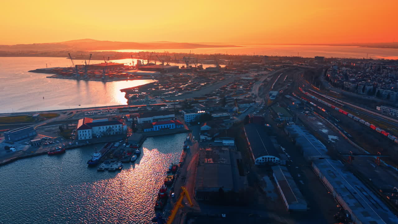 Varm solnedgångsglöd över en storskalig industriell hamnanläggning. Flygfoto på hög höjd över en lasthamn och stad under en livfull orange solnedgång.