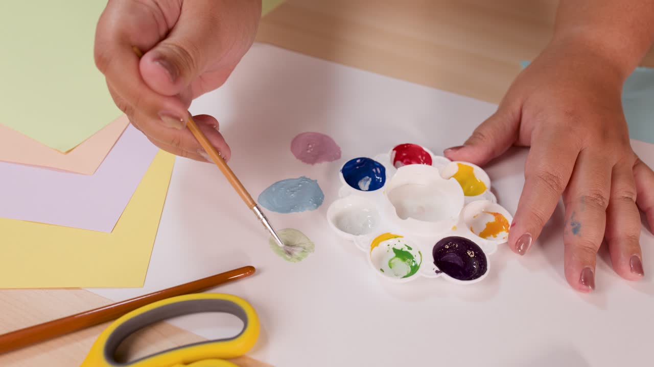 Person mixes watercolor paints with brush on palette, pastel paper background, soft natural lighting