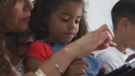 Granddaughter-With-Grandmother-Sitting-On-Sofa-At-Home-Using-Digital-Tablet