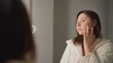 lady uses cosmetic to face standing at mirror. brunette lady with short wet hair in plush robe applies moisturizing gel to face in light place