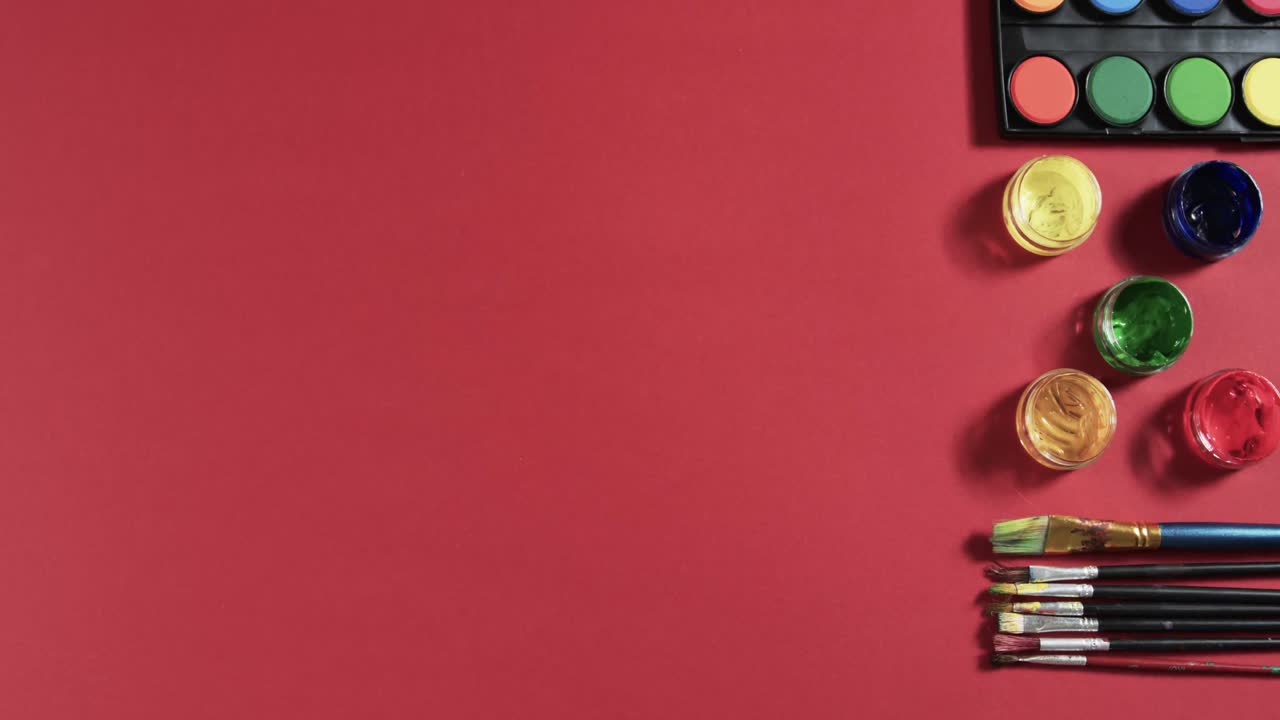 vista aérea de pinturas y pinceles dispuestos sobre un fondo rojo con espacio de copia, en cámara lenta