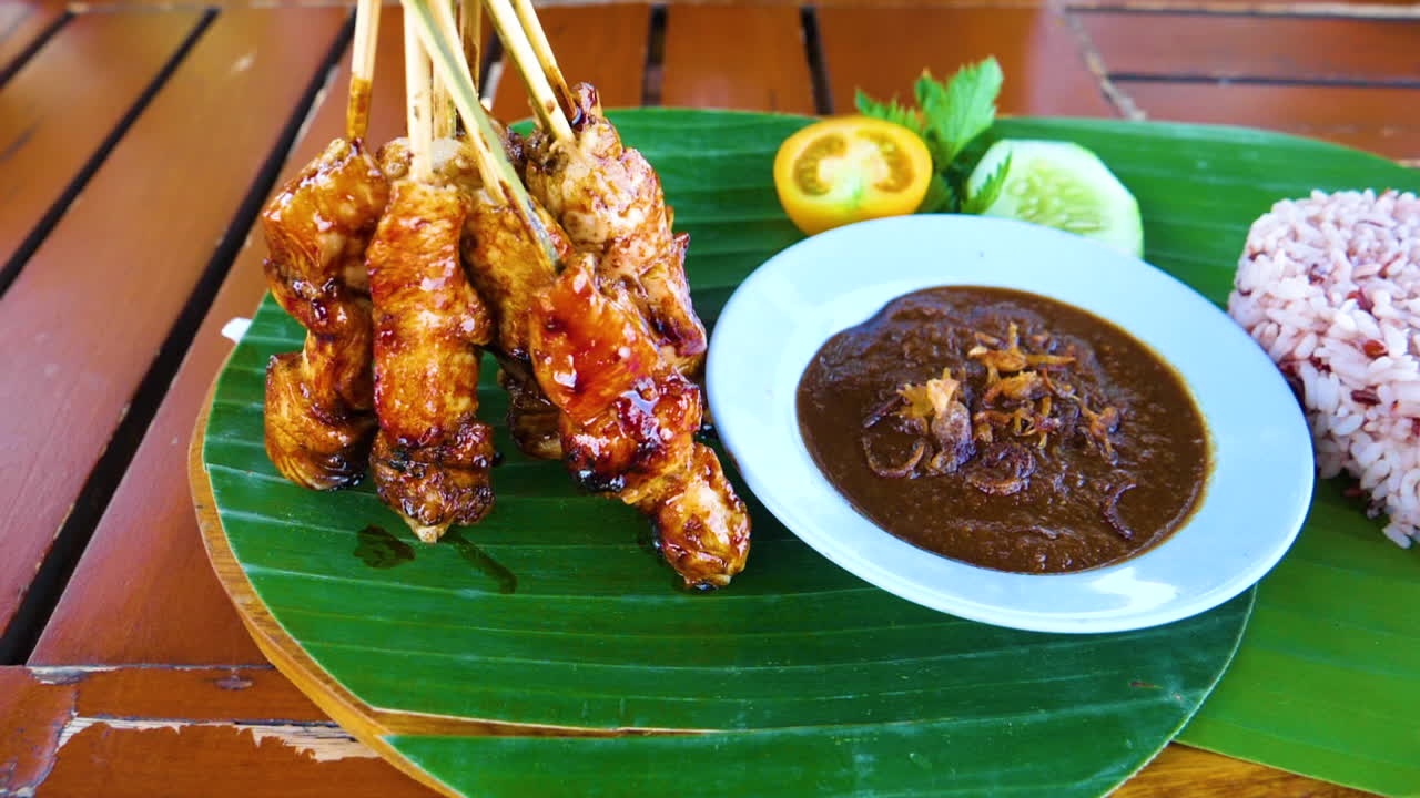 Kana satay, kastike ja sydämenmuotoinen riisi tarjoiltuna vihreillä lehdillä puupöydällä.