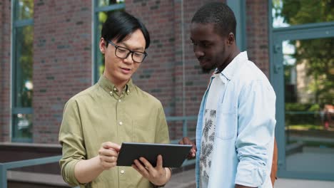 Camera-zooming-on-asian-man-talking-with-his-caucasian-african-american-classmate-watching-something-on-the-tablet-and-talking-in-the-street-near-the-college