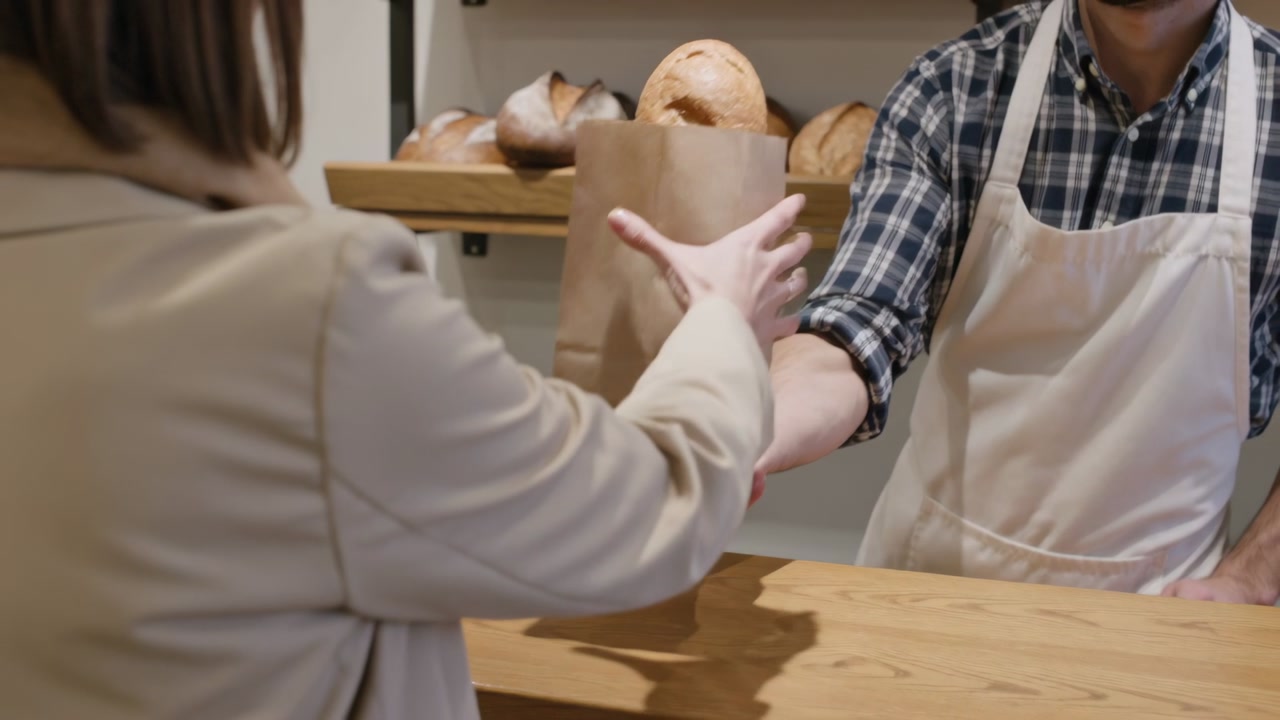 Bagging aproned baker handing loaf in brown paper bag over wooden counter to customer buying bread