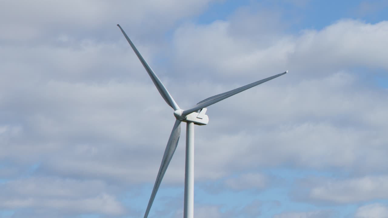 Single wind turbine spins steadily outdoors, daylight, wide shot, partly cloudy sky, static camera