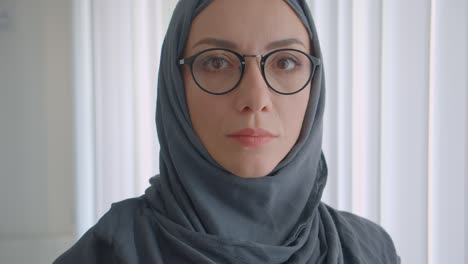 closeup portrait of young attractive muslim businesswoman in glasses and hijab looking at camera smiling cheerfully in the white apartment indoors