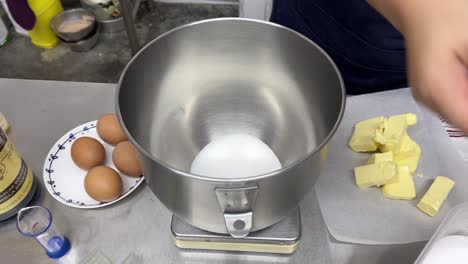 Measuring-precise-quantity-of-caster-sugar-for-baking-recipe,-pouring-into-the-mixing-bowl-on-top-of-the-kitchen-scale-using-measuring-cup,-tilt-down-close-up-shot-at-commercial-kitchen-setting