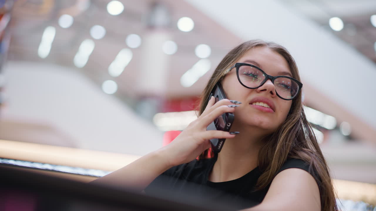 hermosa mujer con camiseta negra y gafas hablando por teléfono, mirando pensativa, con fondo borroso con efecto de luz bokeh y estructuras modernas
