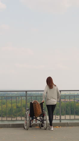 redhead woman stands near wheelchair with little girl. mother and daughter with disability look at river and sand beach from observation deck backside view