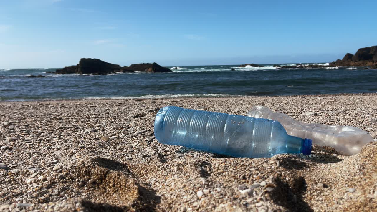 플라스틱 병은 바다의 파도와 멀리 있는 바위 근처의 모래 해변을 오염시니다.