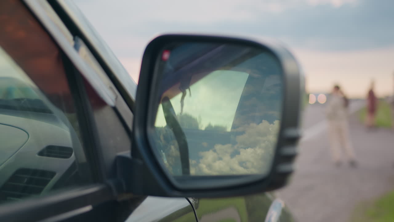 Close up of car side mirror capturing vivid sky reflection while people walk near roadside and distant vehicle with headlights on approaches in soft blur during calm outdoor scene under cloudy sky