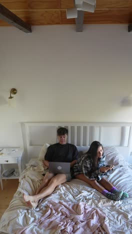 couple relaxing in bed, using laptop and smartphones