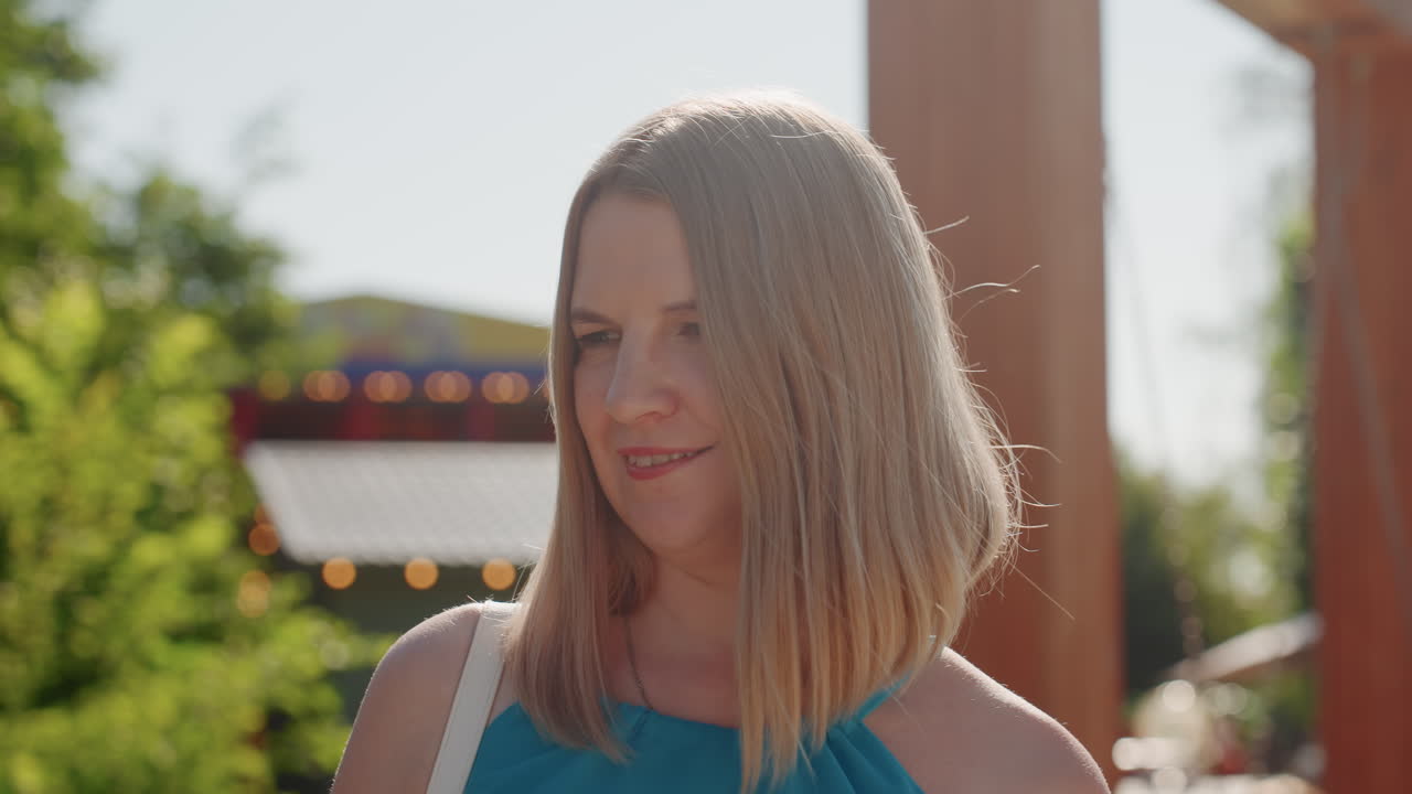 Ritratto in un parco illuminato dal sole di una donna bianca sorridente, scena rilassata all'aperto vicino a un padiglione di legno e catene di luci, capelli biondi all'altezza delle spalle, vestito blu, alone luminoso morbido dal controluce, profondità di campo ridotta, spontaneo.