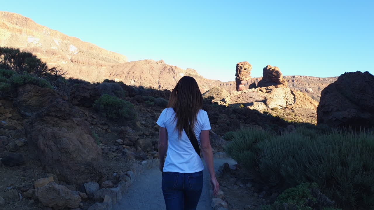 Solitary female tourist walking along scenic trail in Teide national park, capturing volcanic landscape of tenerife with slow motion cinematography under bright sunlight and blue sky