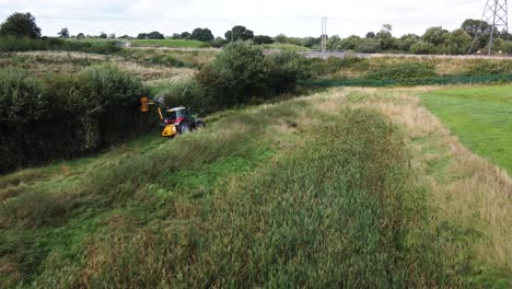 tractor in field trimming, cutting overgrown hedge and bush, follow