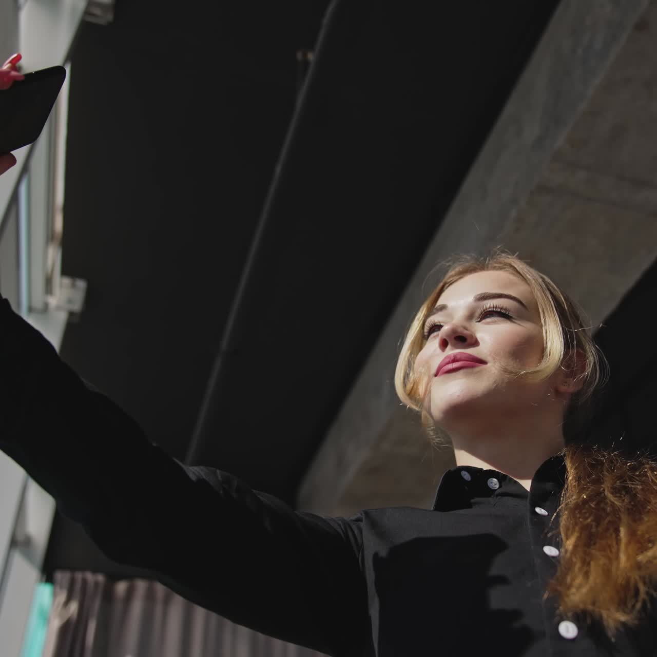 Beautiful lady taking video on her smartphone. Taking selfie near the sunny panoramic window. Low angle view