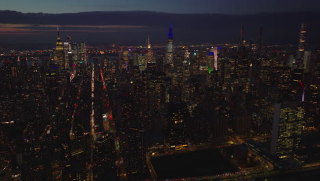 Vista-Aérea-De-Los-Rascacielos-Del-Centro-De-La-Ciudad-Por-La-Noche.-Revelan-Al-Revés-De-La-Calle-Muy-Transitada-En-La-Zona-Ribereña.-Manhattan,-Ciudad-De-Nueva-York,-Estados-Unidos