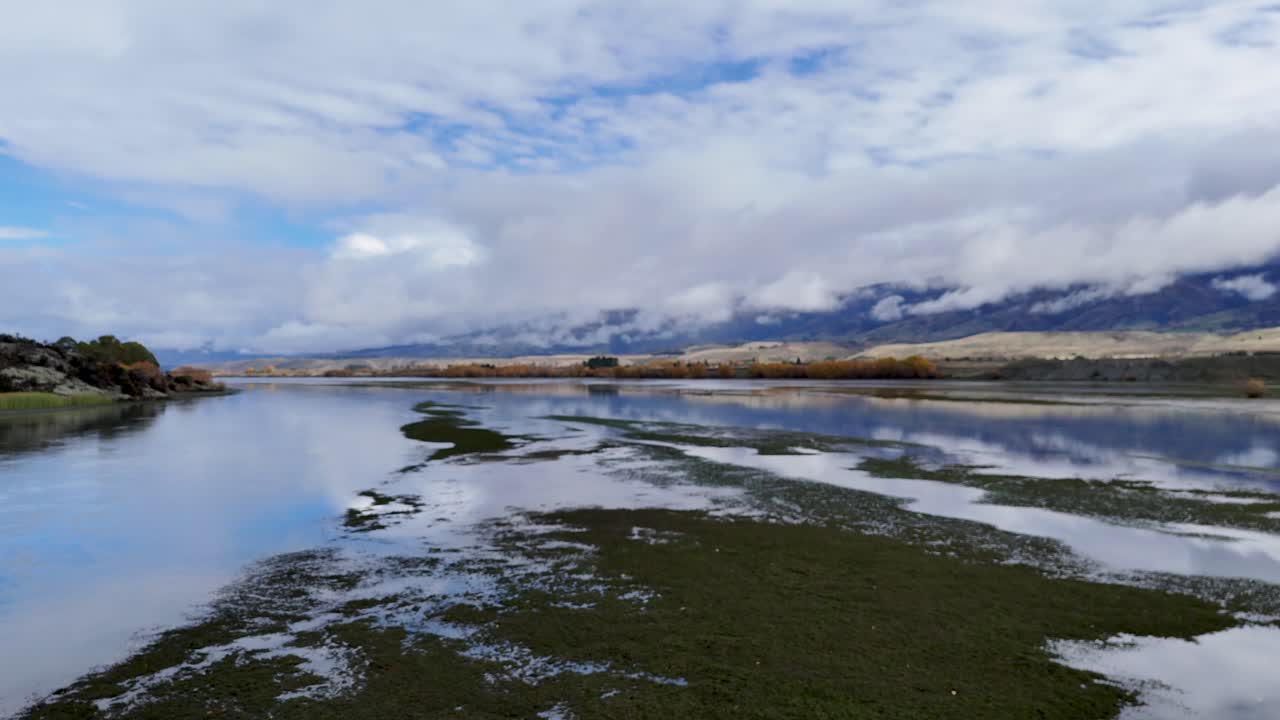 Drone glides above tranquil wetlands with water reflections, lush greenery, and distant mountains under dramatic clouds. Smooth forward camera movement, natural daylight