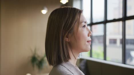 Asian-businesswoman-standing-looking-out-of-window-in-modern-office