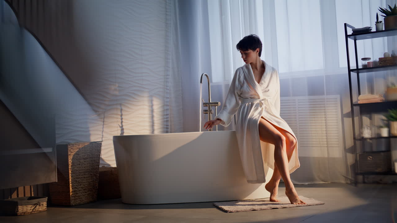 Serene brunette sitting bathtub checking water temperature soft sunlight room