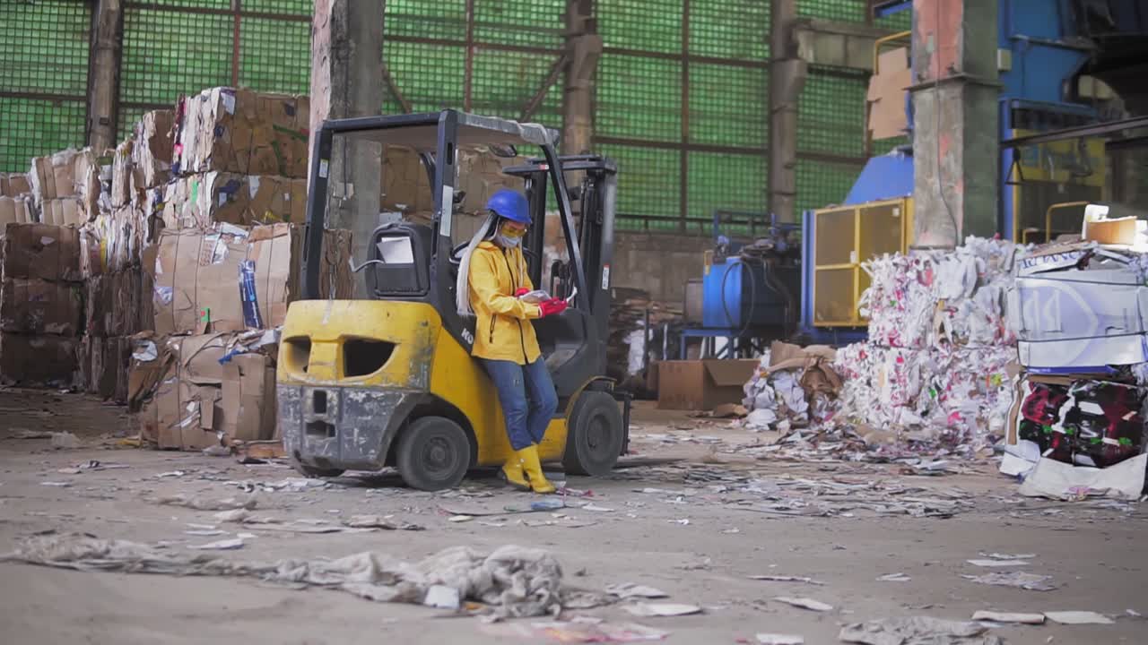 Female worker in hard hat and yellow jacket standing, leaning on warehouse electric car and reading work journal. Huge stocks of pressed carton. Full length