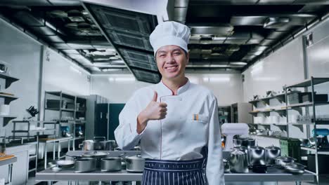 chef asiático sonriendo y mostrando los pulgares hacia arriba gesto a la cámara mientras está de pie en la cocina