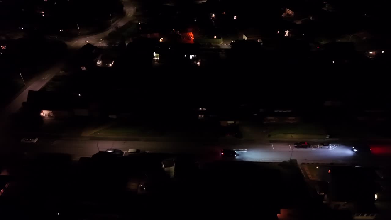 Vista aerea notturna di un'auto che percorre una strada di periferia in America, in una prospettiva di tracciamento, con i fari che illuminano la strada, gli incroci vicini e le aree residenziali circostanti. Campo lungo