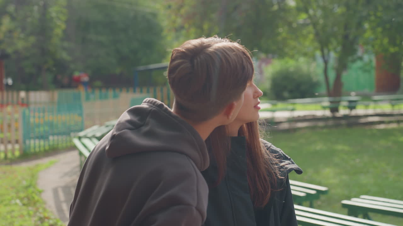 two white teens sitting on bench, summer campmates sharing quiet conversation and soft smiles, boy looking down then meeting girls gaze, green fence and picnic tables nearby, warm sunlight bathing
