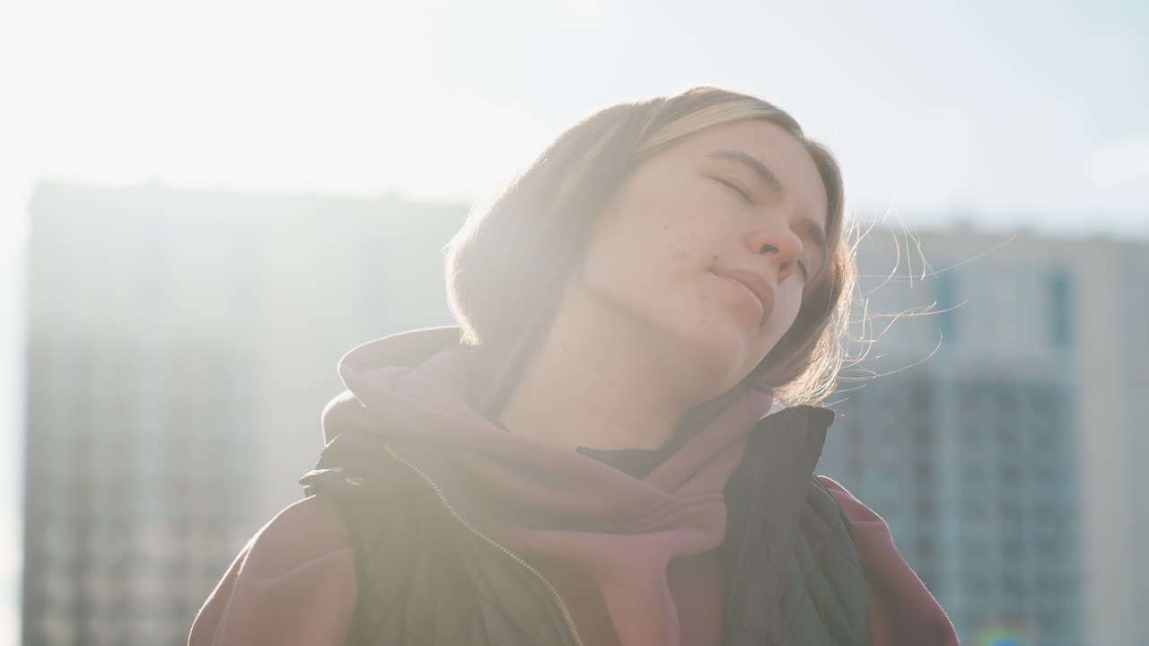 Elegant young lady in hoodie with eyes closed stretches neck with relief while warming up outdoors in urban environment with sunlight highlighting hair and serene expression before workout routine