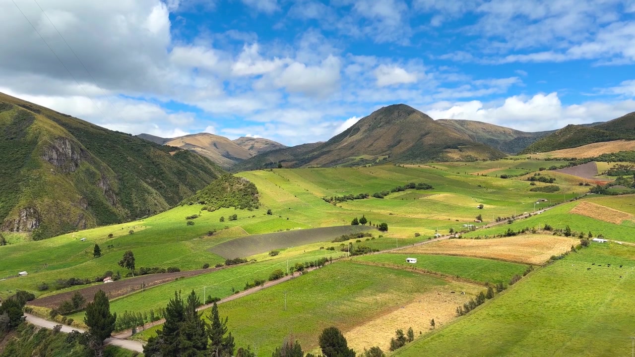 Ampia inquadratura a mano di una strada sterrata che si snoda tra campi agricoli verdi in una soleggiata valle andina con montagne e cielo blu in Ecuador.