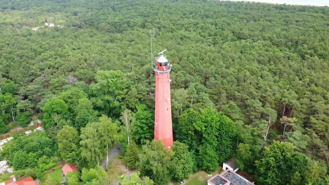 Aerial pullback overview of Hel Lighthouse in Poland, red tower and the surrounding lush green forest along the coast