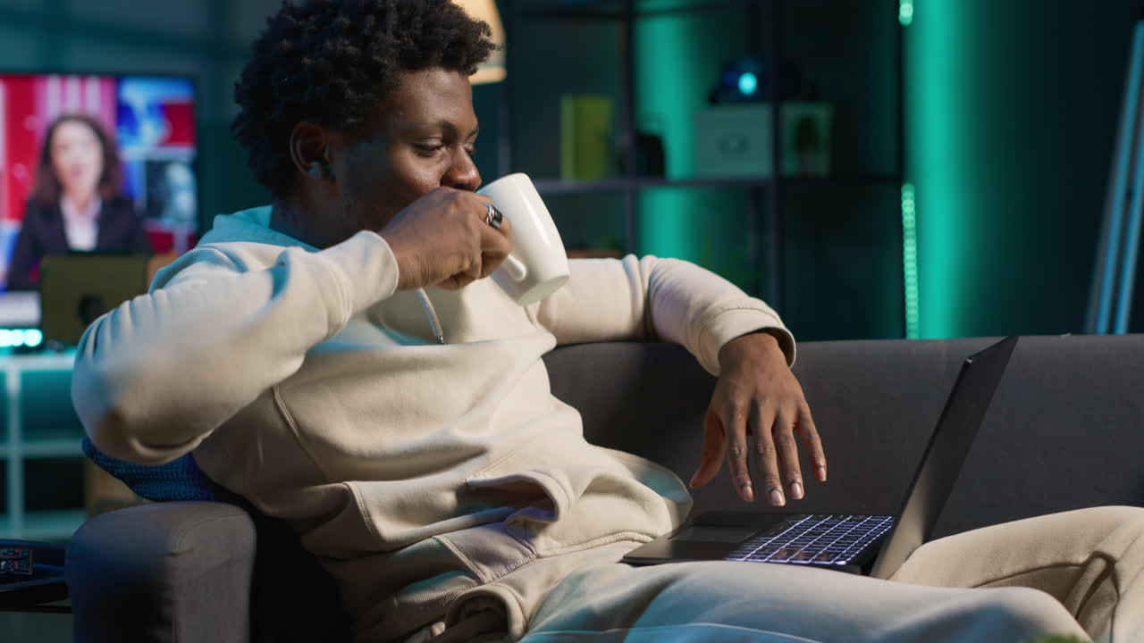 African american person navigating the internet on laptop at his modern luxury loft, relaxing on the couch and browsing web. Cheerful man does freelance work from home. Camera A.