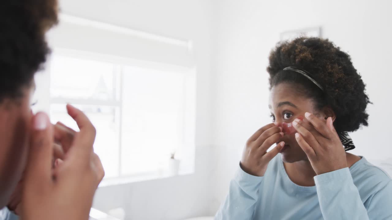 Happy african american woman applying under eye mask looking in bathroom mirror, slow motion