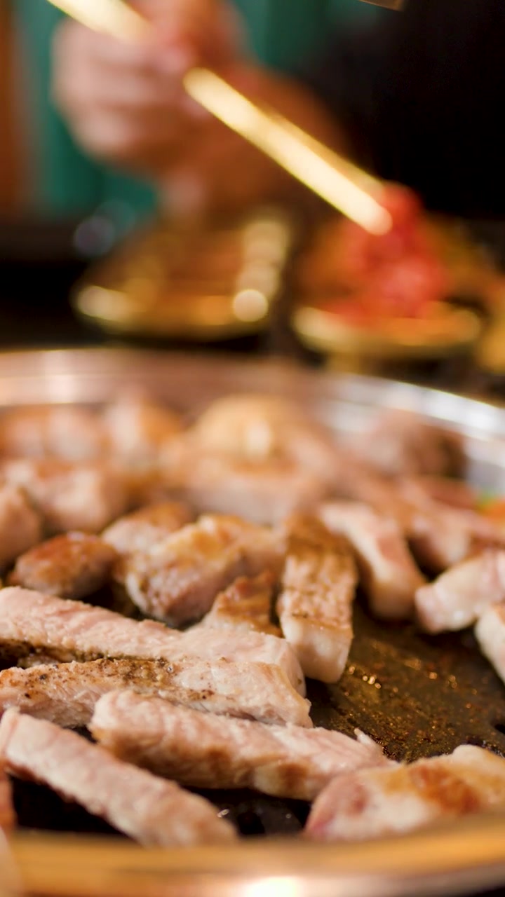 Close-up daging babi dan ayam yang dipanggang di atas hotplate meja, seseorang menggunakan penjepit, pencahayaan hangat.