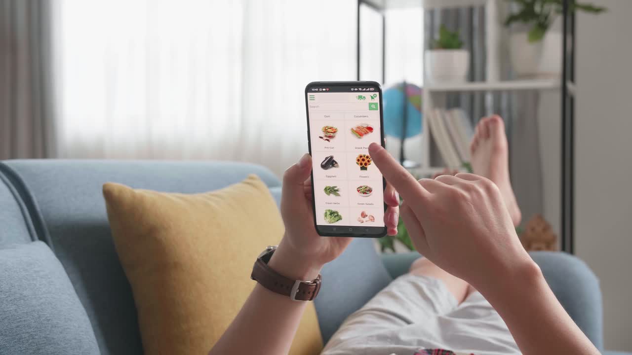 Man Lying On Couch Using Smartphone Buys In Internet Shop. Male Selects Vegetables In Grocery Online Store