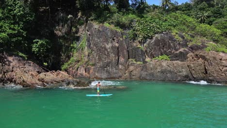 retreat reveal aerial drone bird's eye view of caucasian man exercising on a sup paddle board in turquoise tropical clear waters, coastline and jungle palm trees in thailand