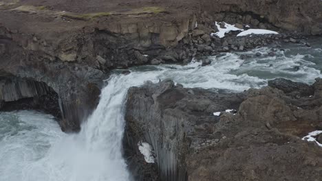 Un-Hombre-Parado-En-El-Borde-Rocoso-Del-Acantilado-Con-Una-Poderosa-Cascada-Que-Fluye-En-Islandia