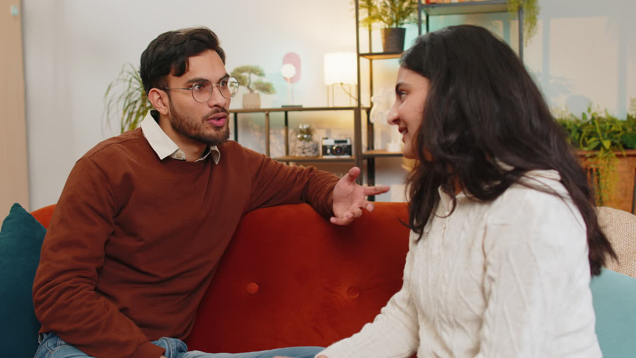 Smiling happy young indian couple sitting on home couch together having lovely conversation relaxing