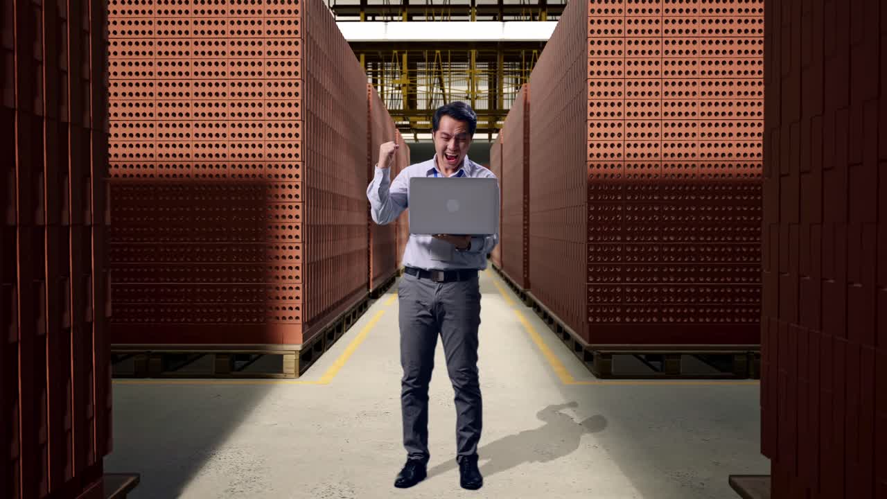 Full Body Of An Asian Male Professional Worker Standing With His Laptop With Red Brick Packed in Stacks Are Stored, He Raises His Fist Up With Screaming Goal