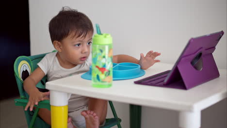 Lindo-Bebé-Latino-Disfrutando-De-Una-Película-En-Una-Tableta-Morada-Mientras-Desayunaba-Sentado-En-Una-Mesa-Blanca-Para-Niños